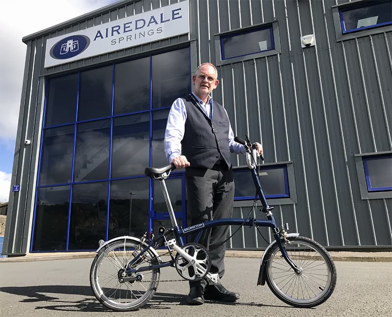 chairman stood infront of Airedale Springs manufacturing facility holding brompton bike