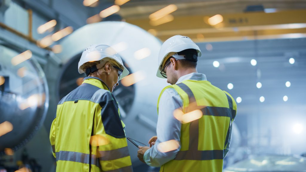 Two Heavy Industry Engineers Stand in Pipe Manufacturing Factory, Use Digital Tablet Computer, Have Discussion. Construction of Oil, Gas and <center>Fuels Transport Pipeline. Back View Sparks Flying