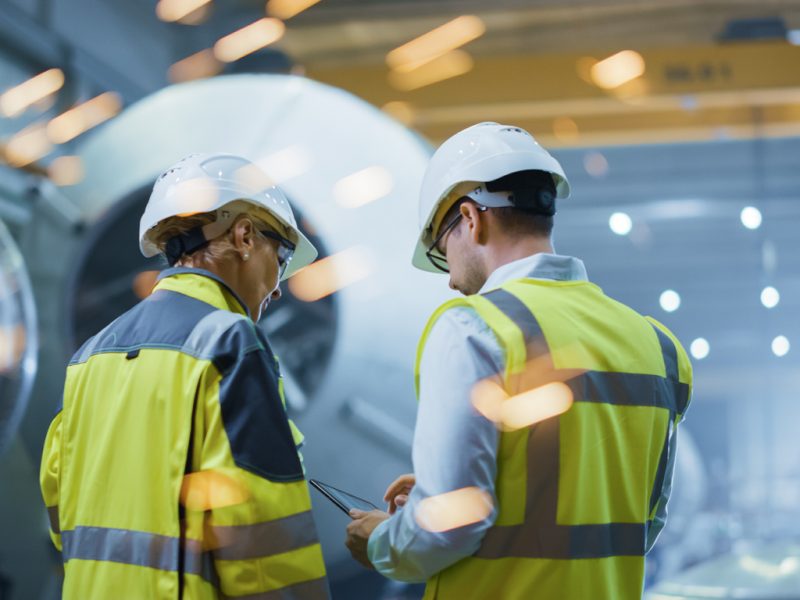 Two Heavy Industry Engineers Stand in Pipe Manufacturing Factory, Use Digital Tablet Computer, Have Discussion. Construction of Oil, Gas and Fuels Transport Pipeline. Back View Sparks Flying